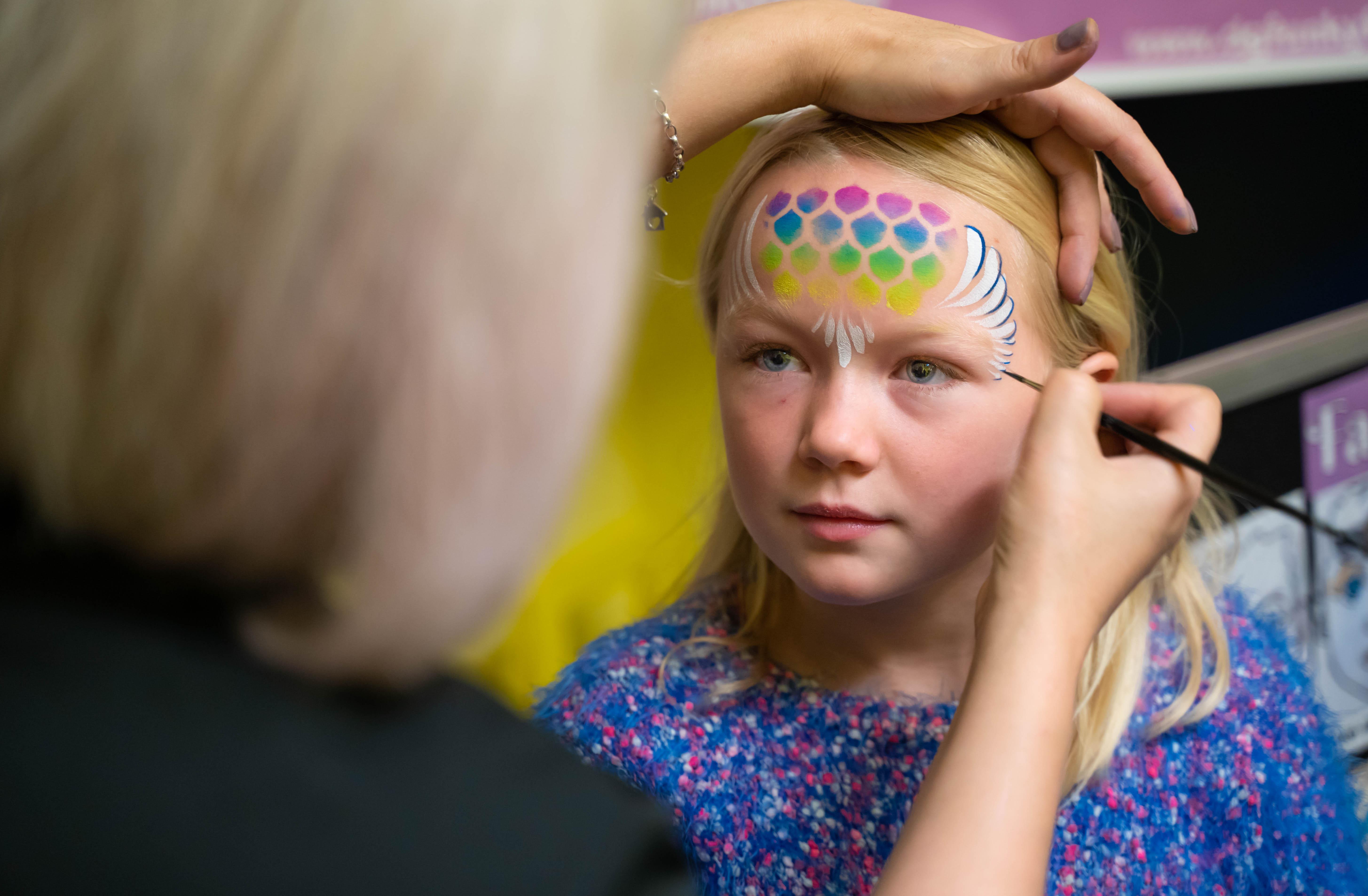 A young child is having her face painted by an adult at a Children's Book Festival event.