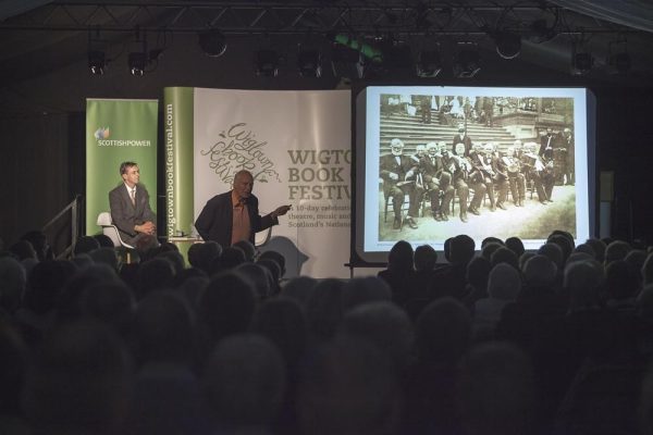 An author talking and showing a slideshow at his event during Wigtown Book Festival. The lights are dimmed, the audience sit listening.
